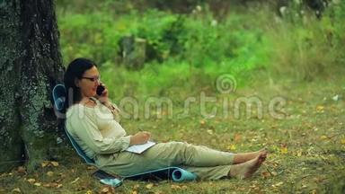 戴着眼镜的微笑着的女人光着脚<strong>坐在</strong>公园的一棵<strong>树下</strong>，用铅笔在笔记本上写字，然后继续说话