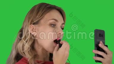 漂亮时尚的年轻女人在嘴唇上涂红色口红，看着绿色屏幕上的手机屏幕，Chroma键。