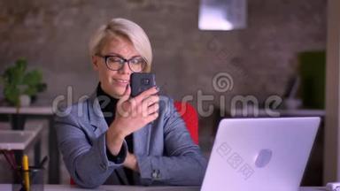 戴眼镜的中年金发短发女商人在办公室里用智能手机在<strong>电玩</strong>上交谈的肖像。