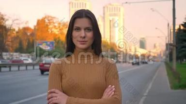 一幅严肃的白种人深色头发的肖像，她用双臂谨慎地注视着镜头，交叉在道路背景上。