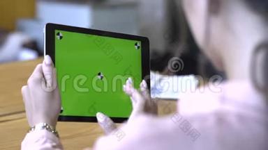 一位棕色头发的年轻女士坐在桌子旁，在彩色钥匙绿色屏幕上滑动着她的平板电脑
