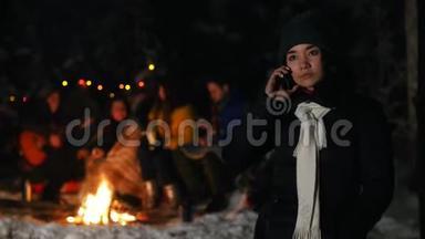 穿着黑色羽绒服的年轻女子站在冬天的森林里，在她的电话里交谈。 一群朋友坐在