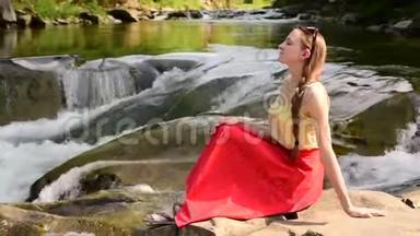 美丽的长发女孩，穿着红色的裙子，坐在山河瀑布背景上的岩石上。 夏日之夜。 安宁