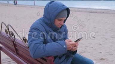 穿着蓝色<strong>羽绒服</strong>的人坐在沙滩上的长凳上，在手机上输入一条信息。