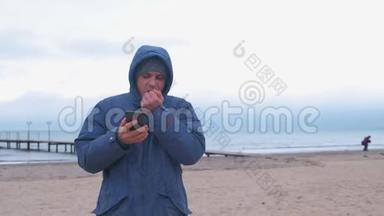 穿着<strong>蓝色羽绒服</strong>的人在海边的沙滩上用冻僵的手在手机上打字。