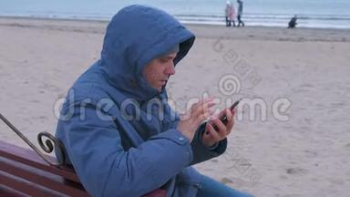 悲伤的男人穿着蓝色<strong>羽绒服</strong>坐在沙滩上的长凳上，打着手机，等待答案。