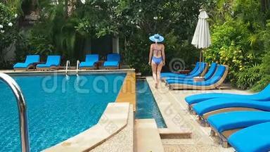 在阳光明媚的日子里，穿着蓝色泳衣、帽子和太阳镜的漂亮女士带着清澈的水在游泳池里走来走去。 奢华