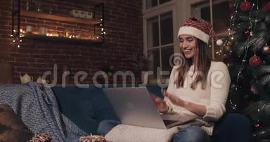 美丽的年轻白种人女孩戴着<strong>圣诞</strong>帽坐在家背景的<strong>圣诞</strong>树附近。 女人用她的笔<strong>记</strong>本电脑