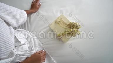 一位穿着<strong>白色浴袍</strong>的年轻女士坐在卧室的床上，她`要打开她男朋友送给她的礼物