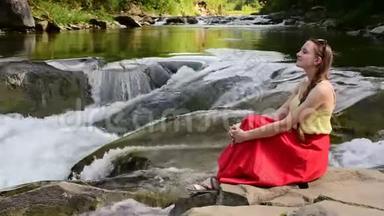 美丽的长发女孩，穿着红色的裙子，坐在山河瀑布背景上的岩石上。 夏日之夜。 <strong>安宁</strong>