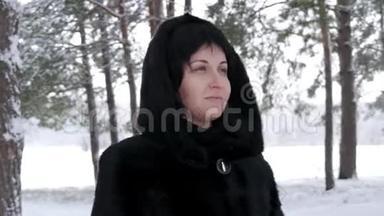 冬天下雪的时候，一个穿着毛皮大衣的黑发女人站在森林或公园里，看着远处。 摄像机移动