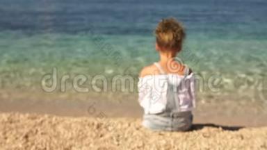 动态视频。 镜头接近坐在美丽清澈大海岸边的女孩.. 女学生转身