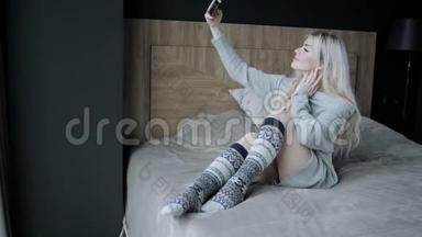 概念<strong>制作</strong>自拍.. 美丽的金发女人坐在床上，带着智能<strong>手机</strong>。 女孩微笑，心情好