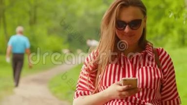孕妇一只手握着电话，快速键入信息，对着镜头微笑，<strong>漫步公园</strong>。 中弹。