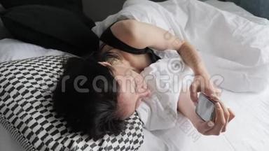 一个幸福的女人躺在她可爱的卧室里，躺在白色的床上，在智能手机上发短信。 技术、互联网