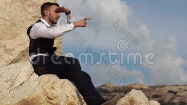 英俊的时尚男士穿着华丽的布坐在岩石上，一边欣赏着令人惊叹的风景，一边向人们展示一些东西
