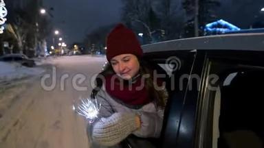 年轻快乐的女人站在车<strong>窗外</strong>，在<strong>下雪</strong>的冬夜玩着火花。 漂亮女孩开车很有趣