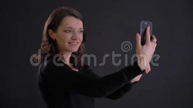年轻迷人的白种人长发女孩微笑着用智能手机在黑色上制作自拍照片的肖像