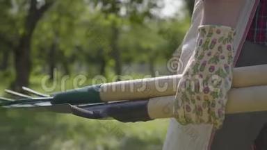 戴着花园手套的农夫手里拿着一把铲子和一把干草叉，在花园里走来走去。 农村概念