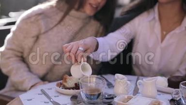 两个漂亮的年轻女人坐在咖啡馆或餐馆的桌子前吃早餐。 女人倒牛奶给她