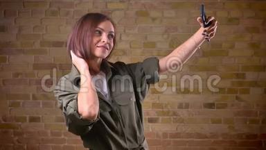 年轻漂亮的棕色头发的女人用智能手机在布里克肯墙的背景下制作自拍照片。