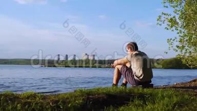 年轻人坐在城市背景的河岸上。 库存录像。 人坐在河边欣赏大自然的景色