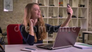 白种人金发女商人在办公室笔记本电脑前的肖像，用智能手机制作自拍照片。