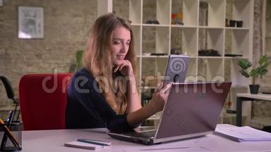 有魅力的金发白种人女商人在办公室笔记本电脑前的肖像，用平板电脑制作自拍照片。
