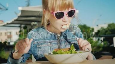 一个戴着粉红色眼镜的女孩正在一家咖啡馆的夏天操场上吃沙拉。 暑假与孩子的概念