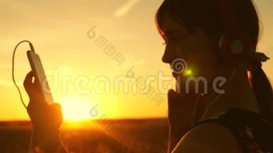 女孩旅行者在智能手机上听音乐，在美丽的春天日落的光线下。 带耳机的年轻女孩
