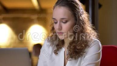 一位年轻的<strong>卷发女士</strong>的特写肖像，她专心地用笔记本电脑工作，在办公室里微笑着进入相机。