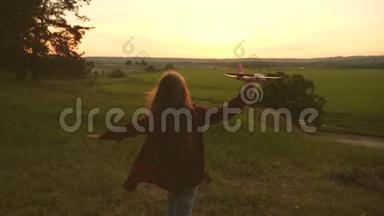 快乐的女孩在日落时带着玩具飞机在田野上奔跑。 快乐童年的概念。 孩子们玩玩具飞机。 少年少年少年