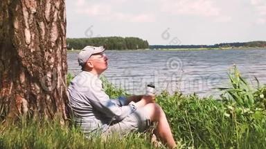 戴帽子的人坐在湖边的一棵树下喝水