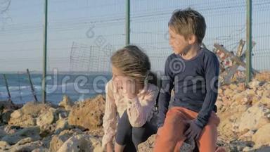 两个可怜的难民儿童男孩女孩坐在海边的石头上。用铁丝网把这个州隔开。