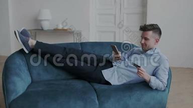 非凡而英俊的男人躺在沙发上，穿着鞋类，舒适地躺在家里的公寓里。 他上网