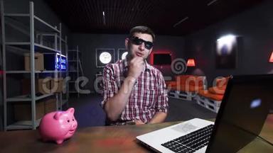 有魅力的年轻人带着太阳镜对着镜头说话，有猪和笔记本电脑在桌子上