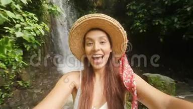 年轻的快乐混合种族旅游博客女孩穿着白衣服和草帽制作自拍视频使用手机在惊人的野生
