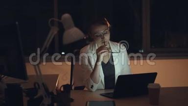 精疲力竭的年轻女士<strong>深夜</strong>在办公室<strong>工作</strong>，用笔记本电脑，然后摘下眼镜，擦脸