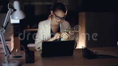 漂亮的金发女郎正在使用智能手机发短信，深夜独自坐在办公室里微笑。 现代小玩意，硬的