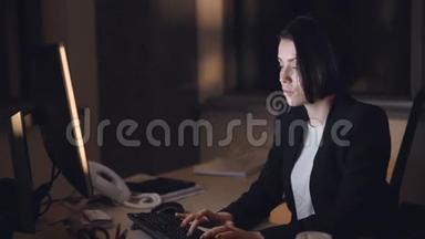 穿着正式西装的女商人正在上电脑夜班，坐在办公桌前打字，看屏幕。 忙碌的年轻人