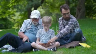 三代幸福的家庭——父亲、祖父和金发儿子坐在公园的草地上，带着书学读书