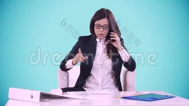 蓝色背景的年轻黑发女人坐在办公室的办公桌后面，在打电话。 桌子上有文件