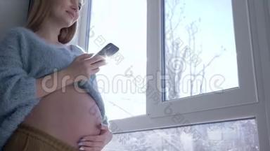 微笑的<strong>大肚</strong>子孕妇在阳光下对着窗户等宝宝的时候使用了现代移动技术