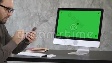 迷人的胡须男检查他的牙齿在他的手机自拍相机附近的电脑显示器。 绿色屏幕模拟显示。