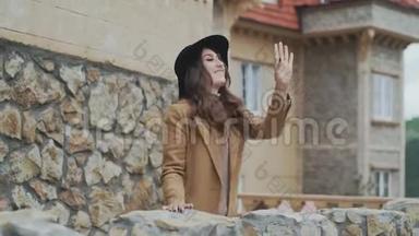 一位戴着帽子的漂亮年轻女子站在一个石质阳台上，挥舞着她的手，和一个陌生人说话