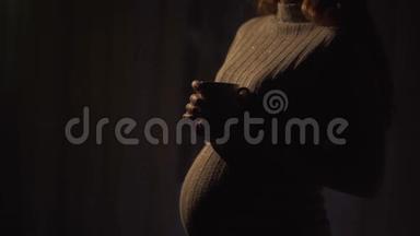 不明身份的怀孕女孩站在温暖的毛衣在黑暗的房间里喝热茶。 女士在等