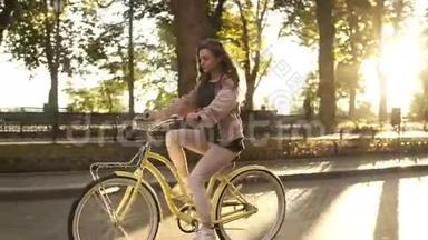 侧景漂亮的长发女孩骑自行车在街道或林荫大道在夏季城市公园。 穿着粉色衣服