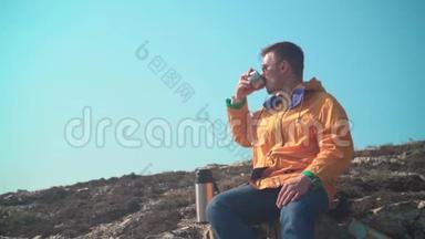 一个穿着黄色夹克，蓝色牛仔裤和眼镜的男人坐在山上，欣赏风景，<strong>喝热水</strong>瓶里的茶。