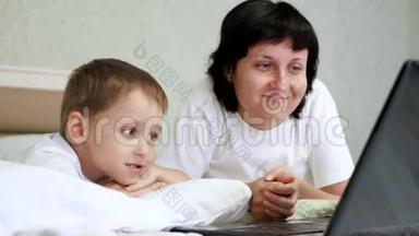 幸福的母亲和她的孩子躺在床上，微笑着看着笔记本电脑屏幕。