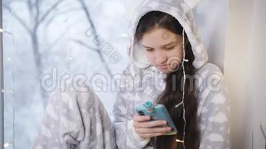 少女冬暖夏凉的睡衣坐在窗边用智能手机.. 窗外下雪了。